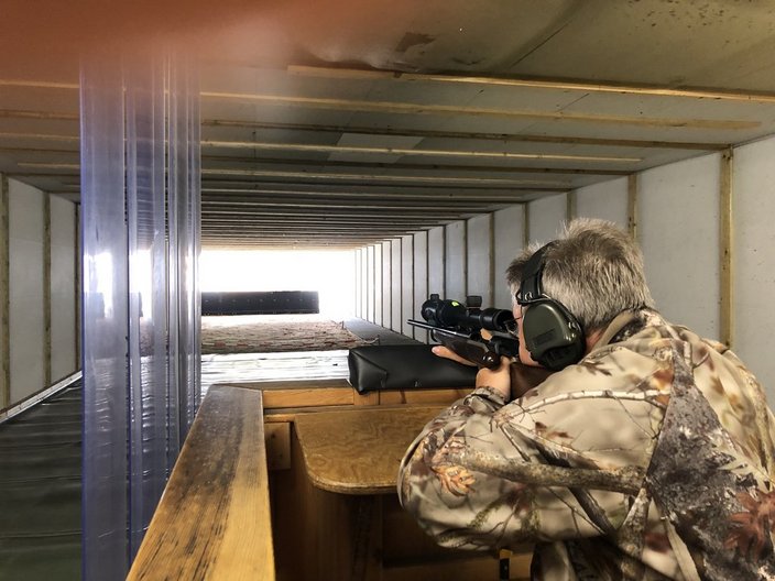 Person zielt mit einem Gewehr, trägt Gehörschutz im geschlossenen, schallgeschützten Schießstand, umgeben von Holzwänden und einer transparenten Trennwand.